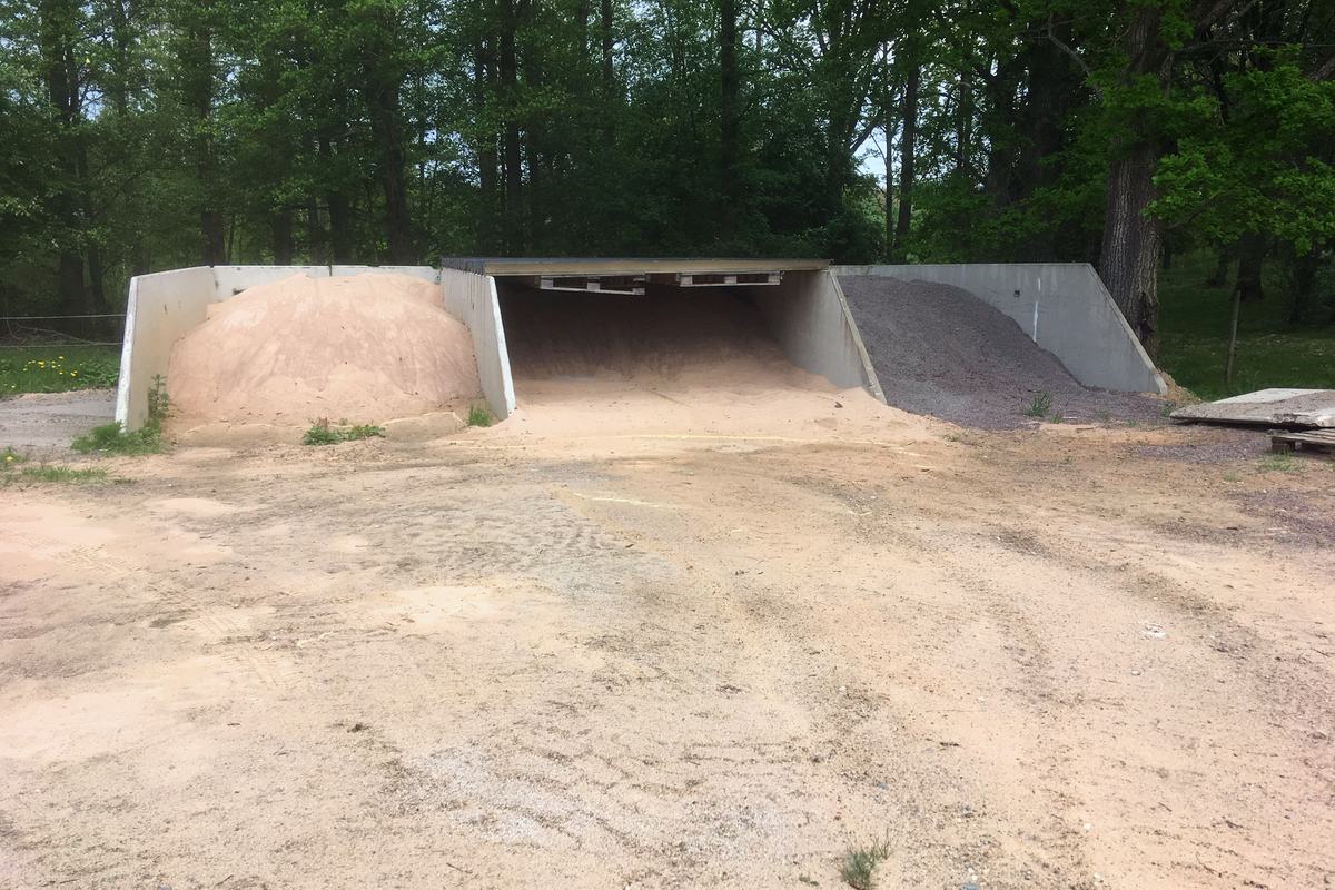 Fack med sand och grus under skärmtak, omgivet av skog.